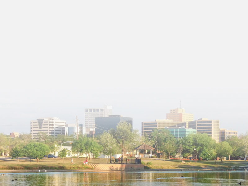 A view of Odessa, TX with buildings in the background and a park with a pond with ducks in it