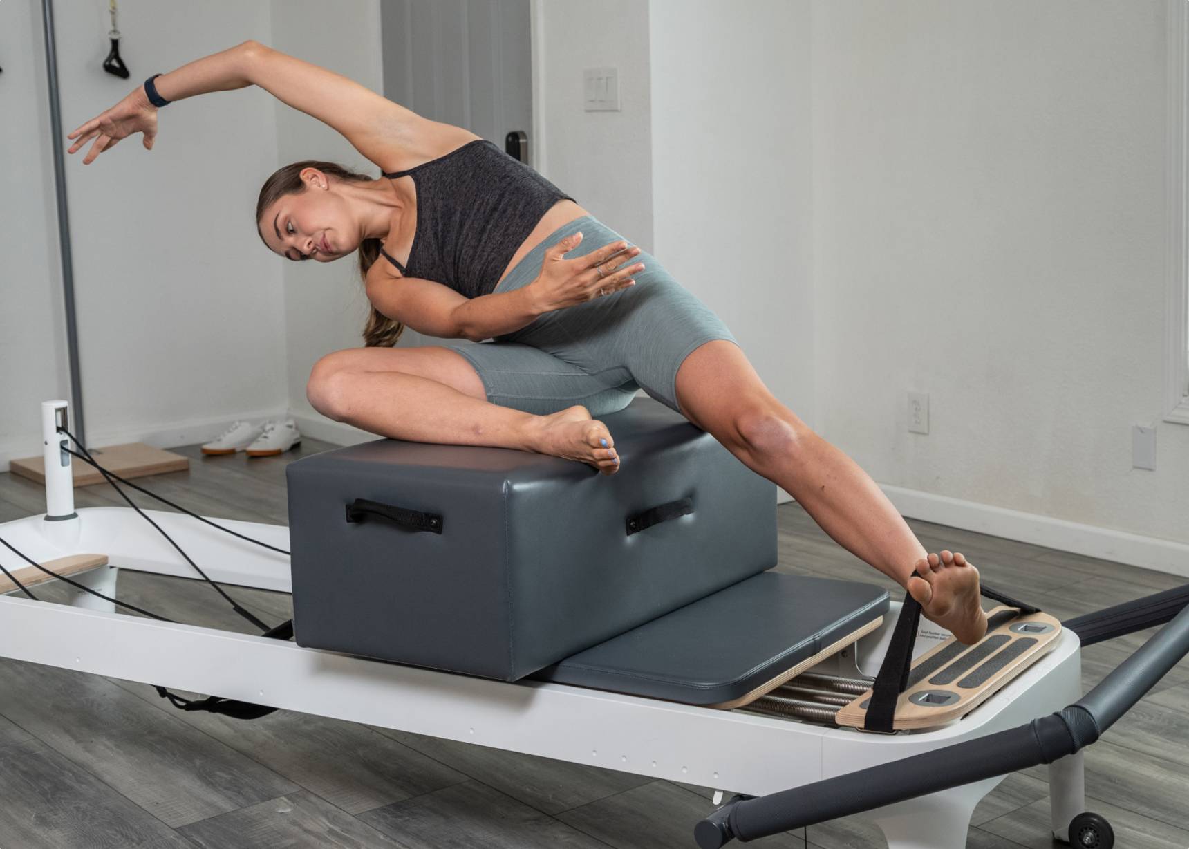 Woman stretching on a sitting box, using Pilates foot straps attached to a reformer.